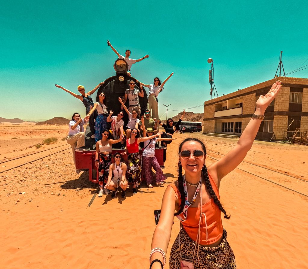 A large group of people poses playfully on and around an old train engine in a vast desert landscape during a weroad group trip.