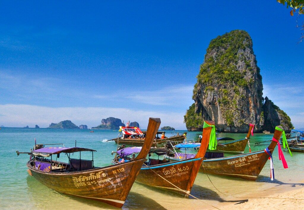 Longtail boats on a sandy beach with a large limestone karst island in the background under a blue sky in Thailand.