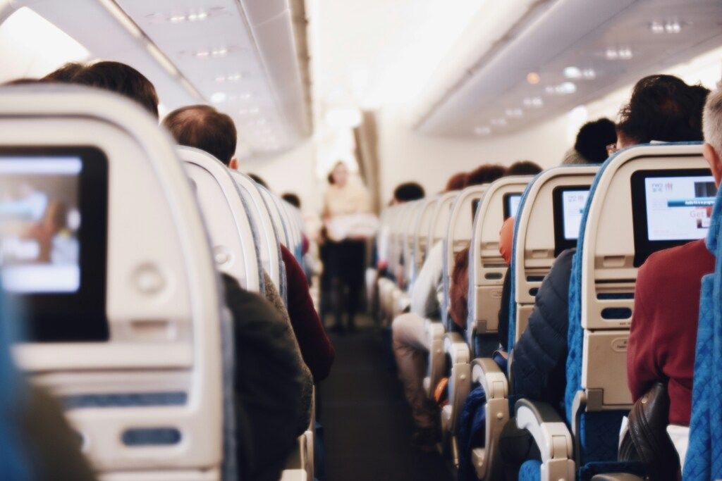 The interior of an airplane cabin, showing rows of passenger seats with screens on the seatbacks and a flight attendant in the aisle.