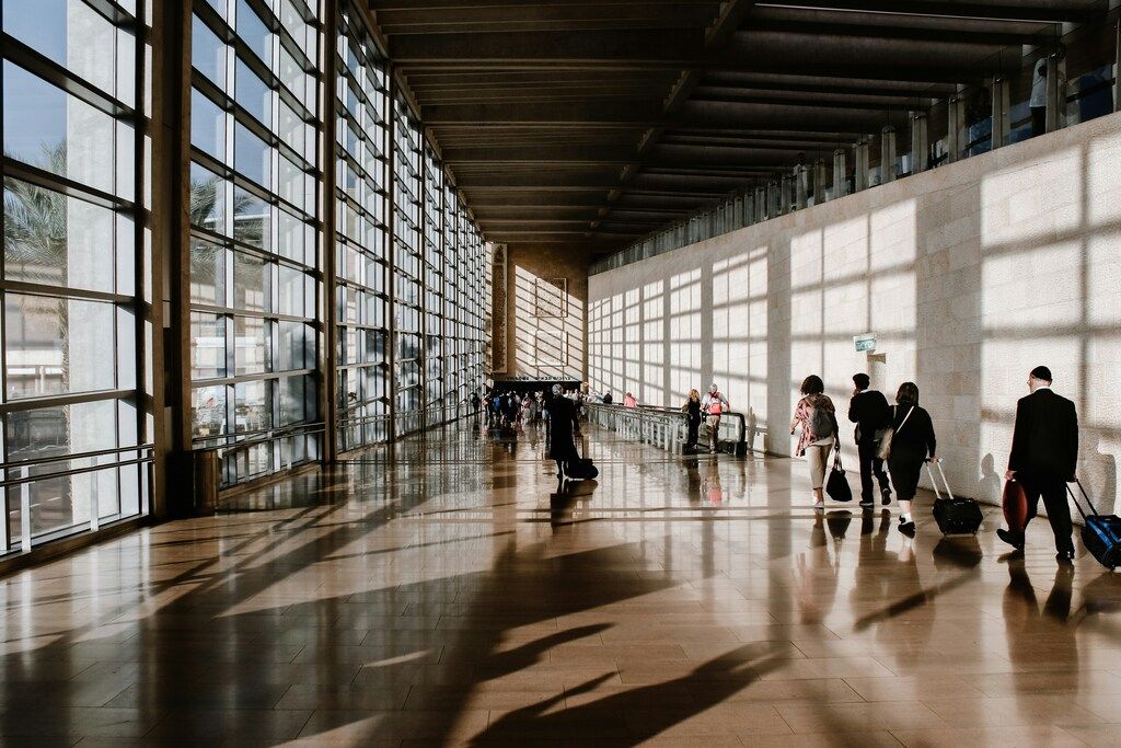 A bright, modern airport terminal hallway with large windows casting long shadows on the polished floor, and several travelers with luggage walking.