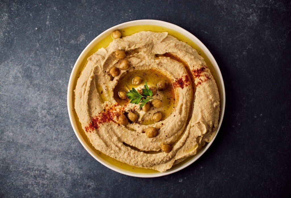 A bowl of hummus garnished with chickpeas, paprika, and a sprig of parsley, drizzled with olive oil.