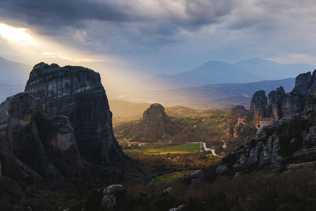 A breathtaking view of the Meteora rock formations and monasteries in Greece, with dramatic cliffs and a valley bathed in sunlight under a cloudy sky.