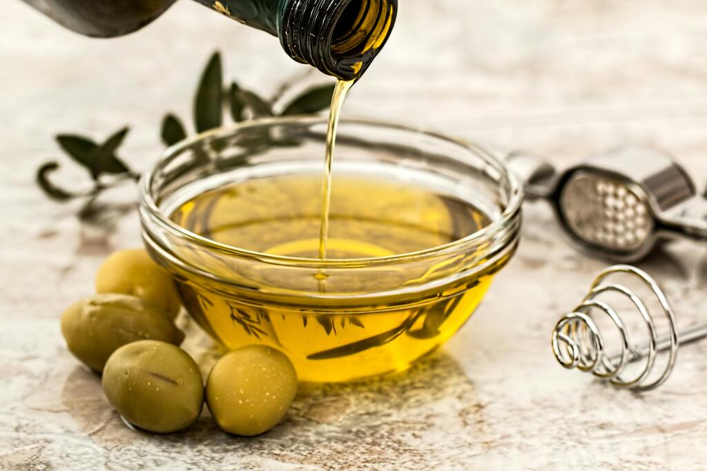 Olive oil being poured from a dark bottle into a clear glass bowl, with green olives and olive branches on a light-colored surface, and a garlic press and whisk in the background.