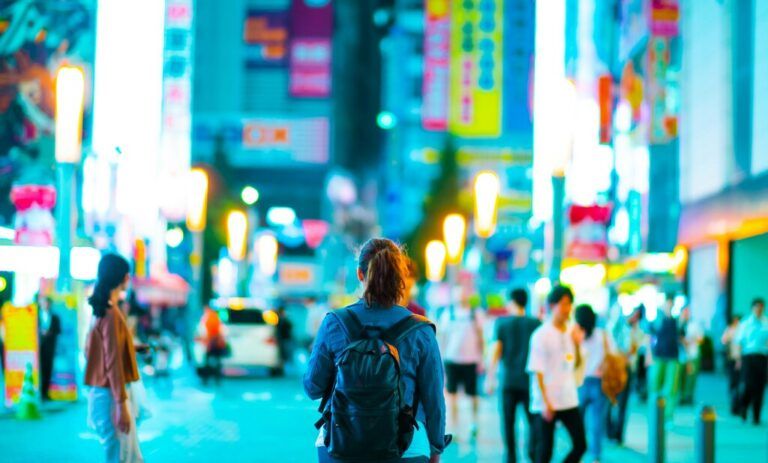 A person with a backpack walks through a vibrant, neon-lit street in a Japanese city, with blurred signs and crowds in the background.