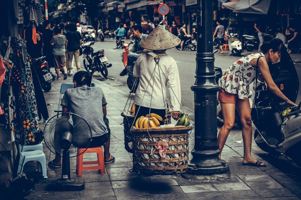 A street vendor wearing a traditional conical hat carries baskets of bananas on a pole along a busy sidewalk in Vietnam.