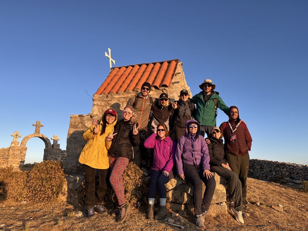Happy WeRoad group posing together by a small stone chapel during a hike on a sunny day.
