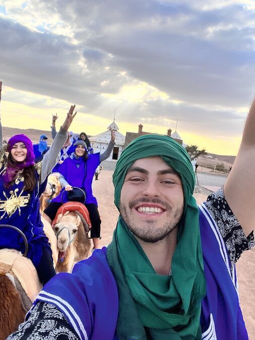 Happy group of WeRoad travelers taking a selfie while riding camels in traditional desert attire at sunset.