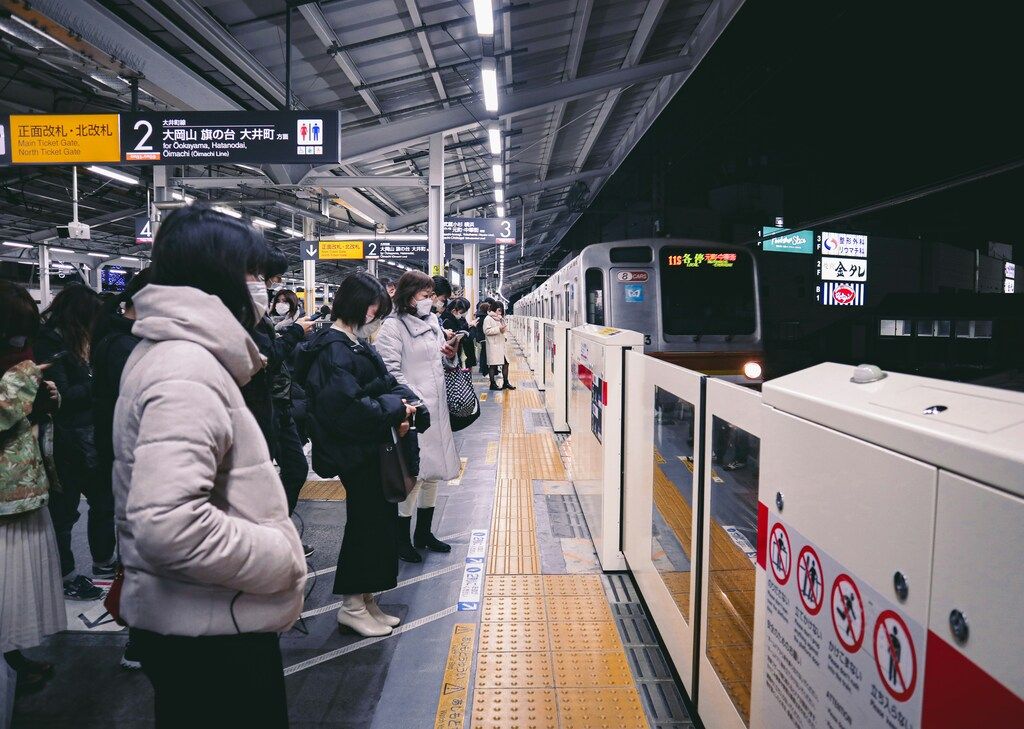 Crowd of passengers waiting for a commuter train at a Japanese station at night, many wearing face masks.