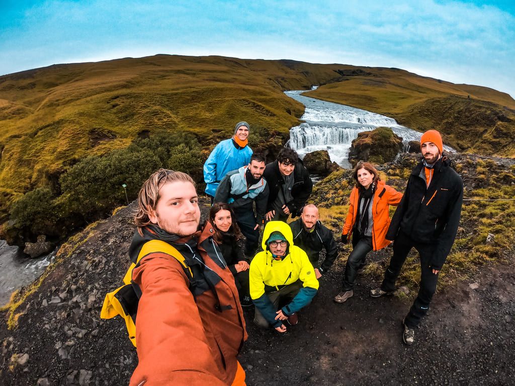 Group of travelers on a WeRoad trip smiling for a selfie in front of a scenic waterfall surrounded by green hills.