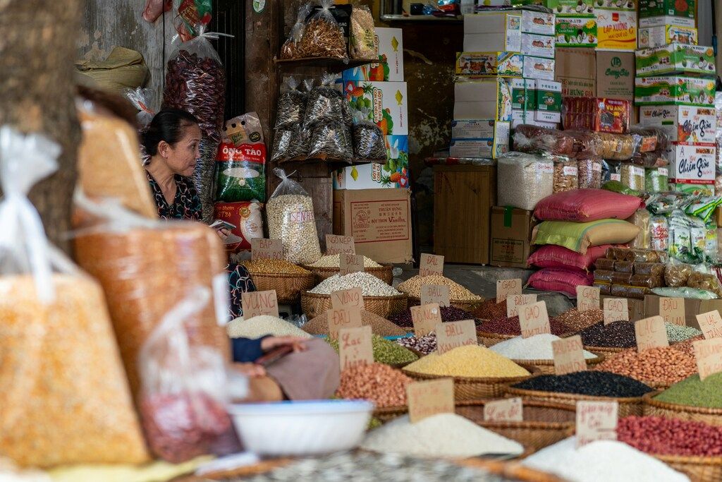 Vietnamese vendor surrounded by baskets of grains, legumes, and spices at a traditional market stall.