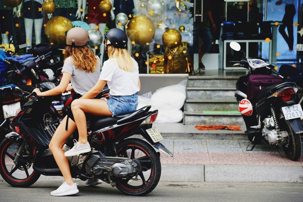 Two young women wearing helmets riding a scooter in a vibrant urban street of Vietnam.