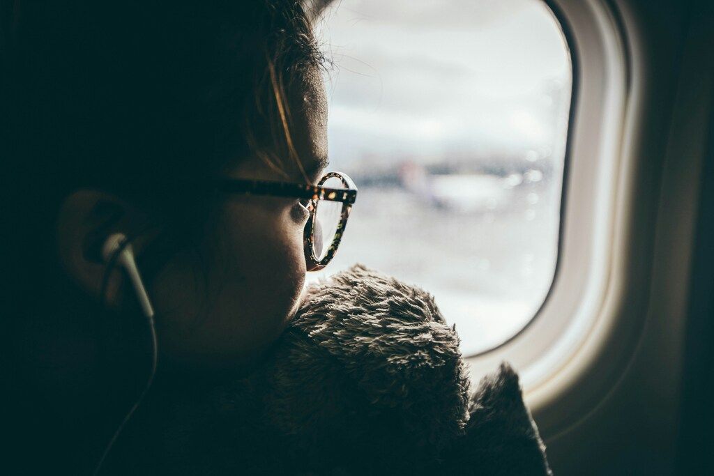A person wearing glasses and earphones, cuddling a plush toy and gazing thoughtfully out of an airplane window.