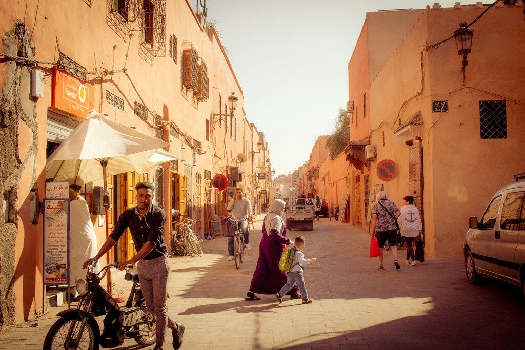 Daily life scene in a sunlit street of Marrakech, with people walking, biking, and shopping.