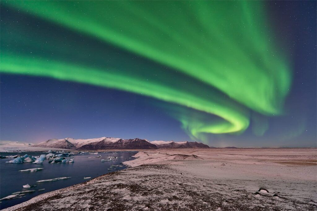 The green Northern Lights swirl in a dark sky above snowy mountains and icebergs floating in a lake.