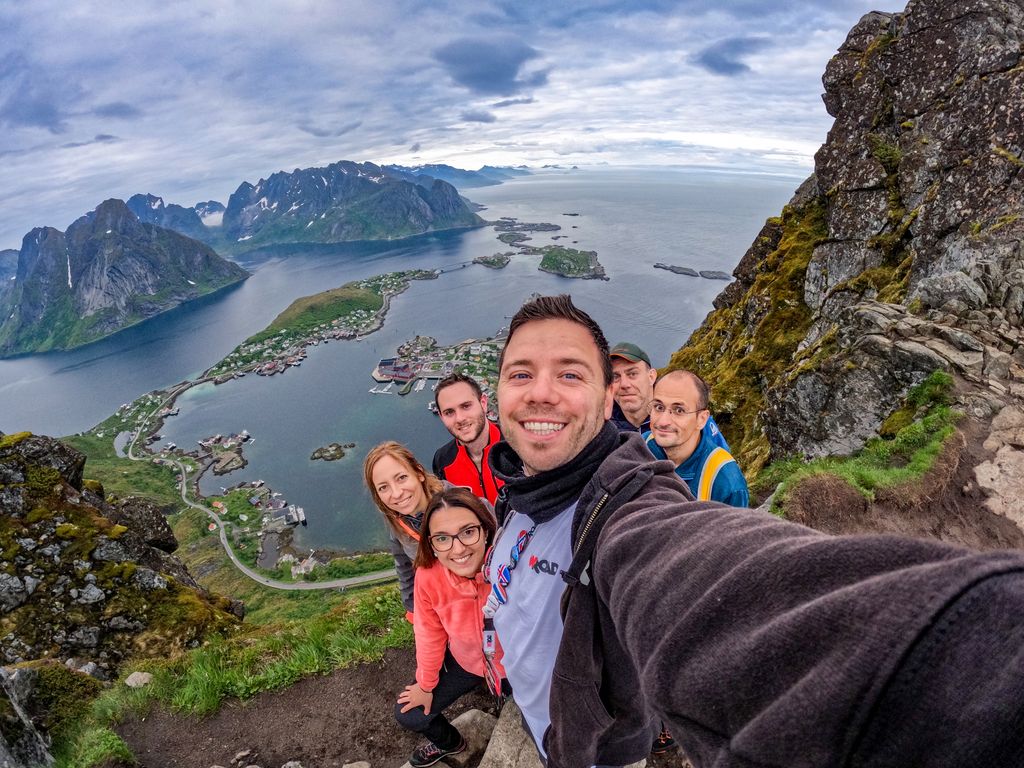 A group of WeRoad travelers taking a wide-angle selfie on a rugged mountain peak overlooking the fjords of the Lofoten Islands.