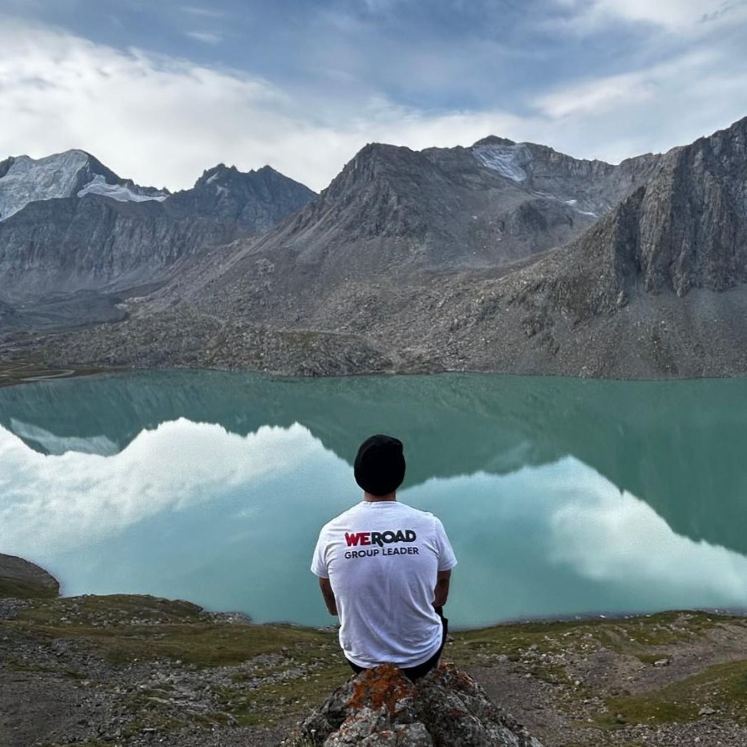 A WeRoad coordinator sitting facing Lake Ala Kul, surrounded by the blue mountains of the Tien Shan in Kyrgyzstan.