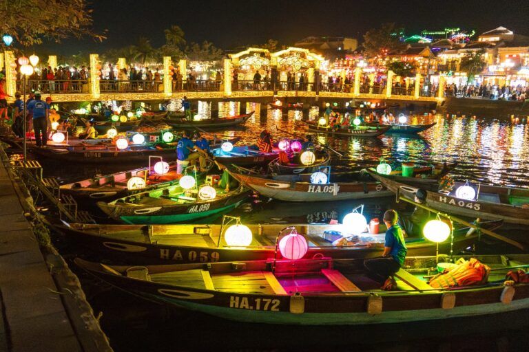Hoi An Lantern Festival at night, showing traditional boats with illuminated lanterns on the river and a festive atmosphere.
