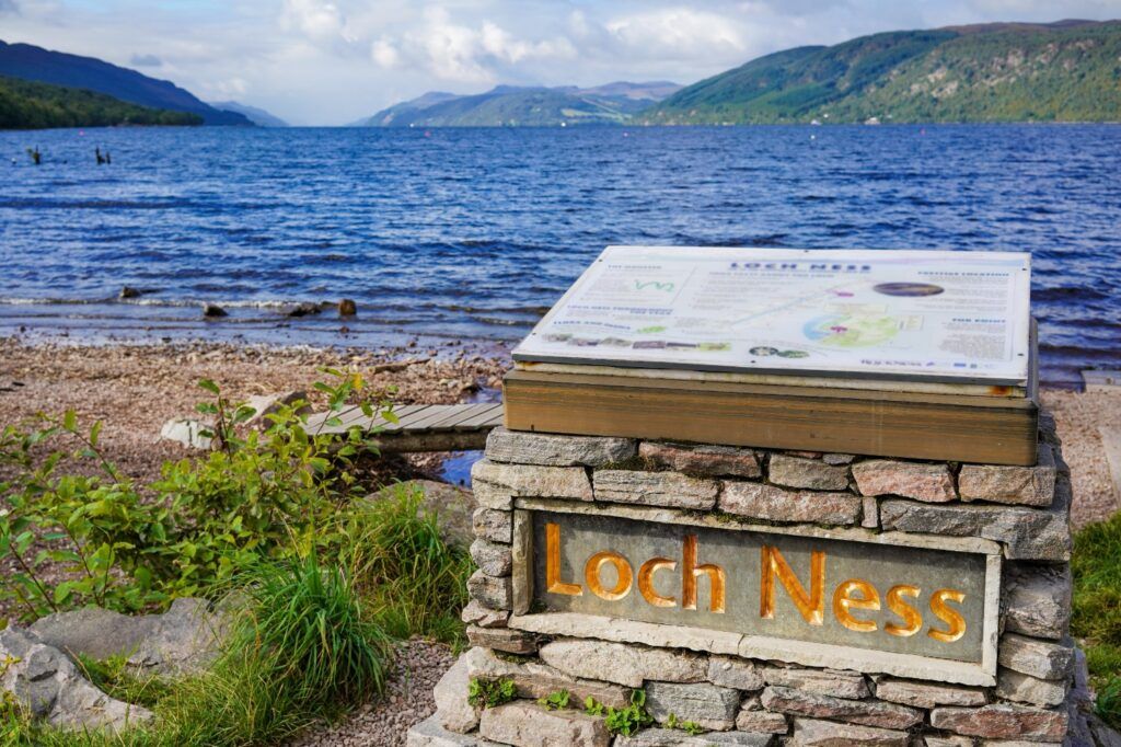 Scenic view of Loch Ness with an information sign on the shore, surrounded by the Scottish Highlands.