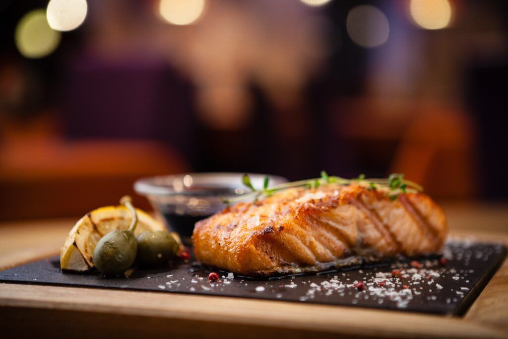 A seared salmon fillet on a slate plate with lemon, olives, and herbs.