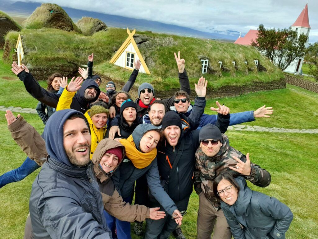 WeRoad travelers taking a selfie in a Scottish landscape with grass-roofed houses and a church.