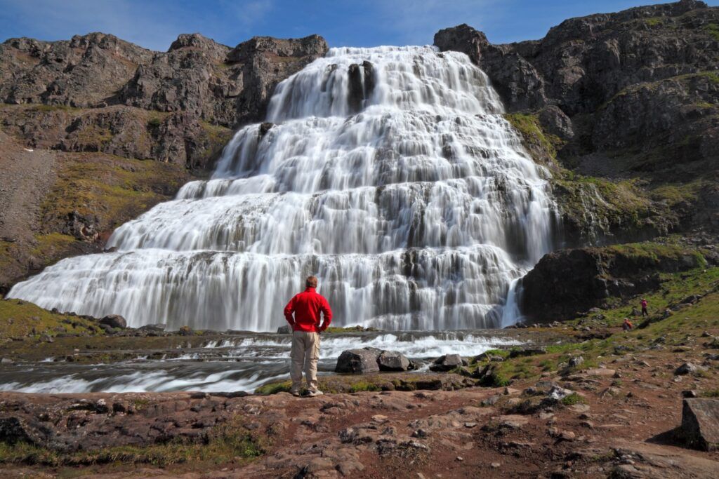 Person in a red shirt standing before the massive, multi-tiered Dynjandi waterfall in Iceland.