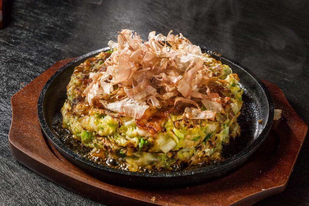 Savory Okonomiyaki (Japanese pancake) served on a hot griddle, topped with sauce and bonito flakes.