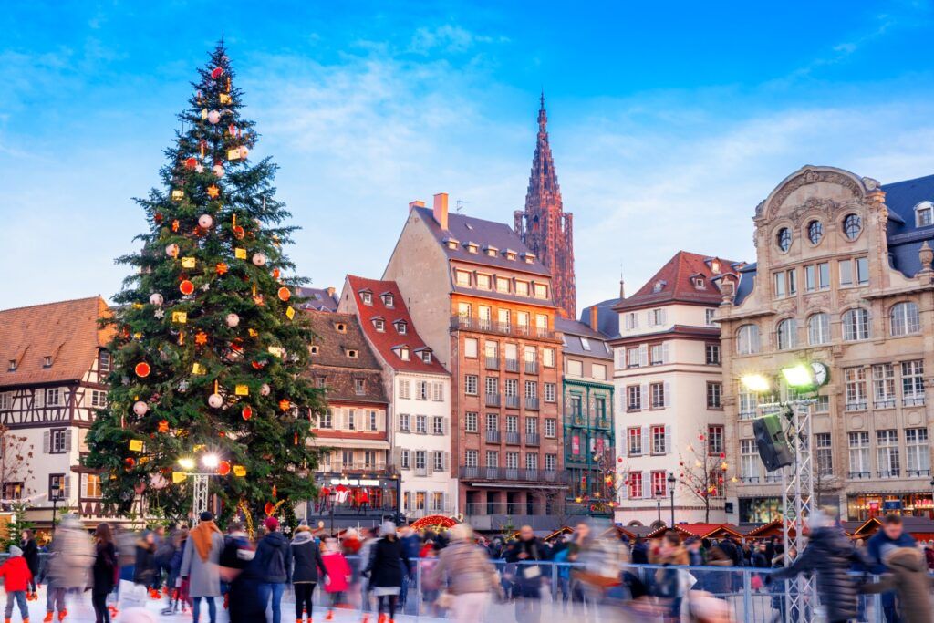 Christmas market scene in Strasbourg with a large decorated Christmas tree, historic timber-framed buildings, and people ice skating.