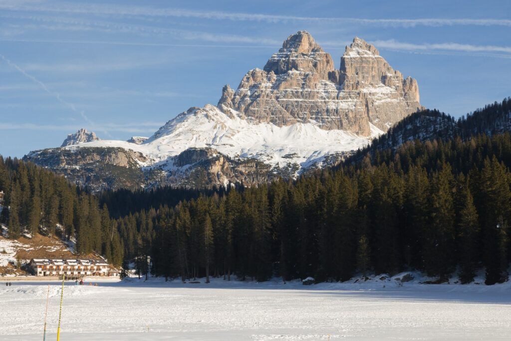 Scenic view of the snow-covered Lavaredo Peaks in the Dolomites, rising above a dark forest and a frozen lake.