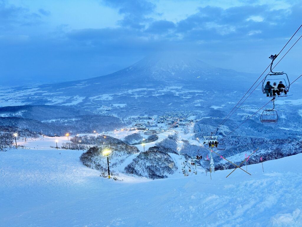 Night skiing in Japan (Japow) with chairlifts ascending towards the illuminated Niseko village and Mount Yotei in the background.