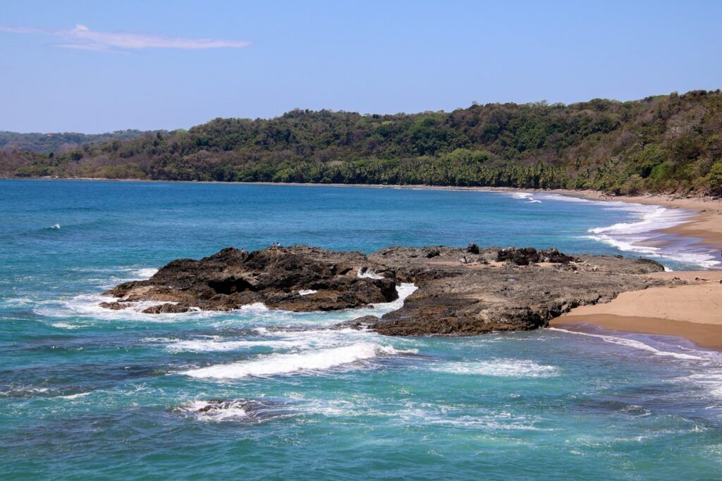 Scenic view of a rocky coastline and beach with turquoise ocean waves and forested hills.