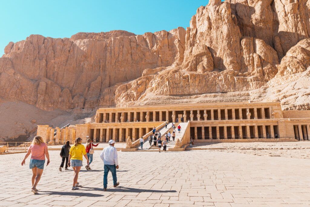 Tourists walking towards the large, columned mortuary Temple of Hatshepsut set against a massive sandstone cliff in Egypt.