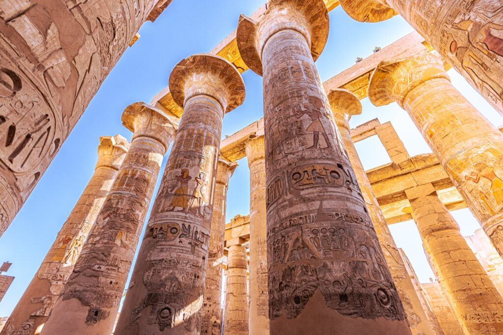 Low-angle view of massive, colorful, inscribed columns with papyrus capitals in the Great Hypostyle Hall of the Karnak Temple, reaching towards the bright blue sky.