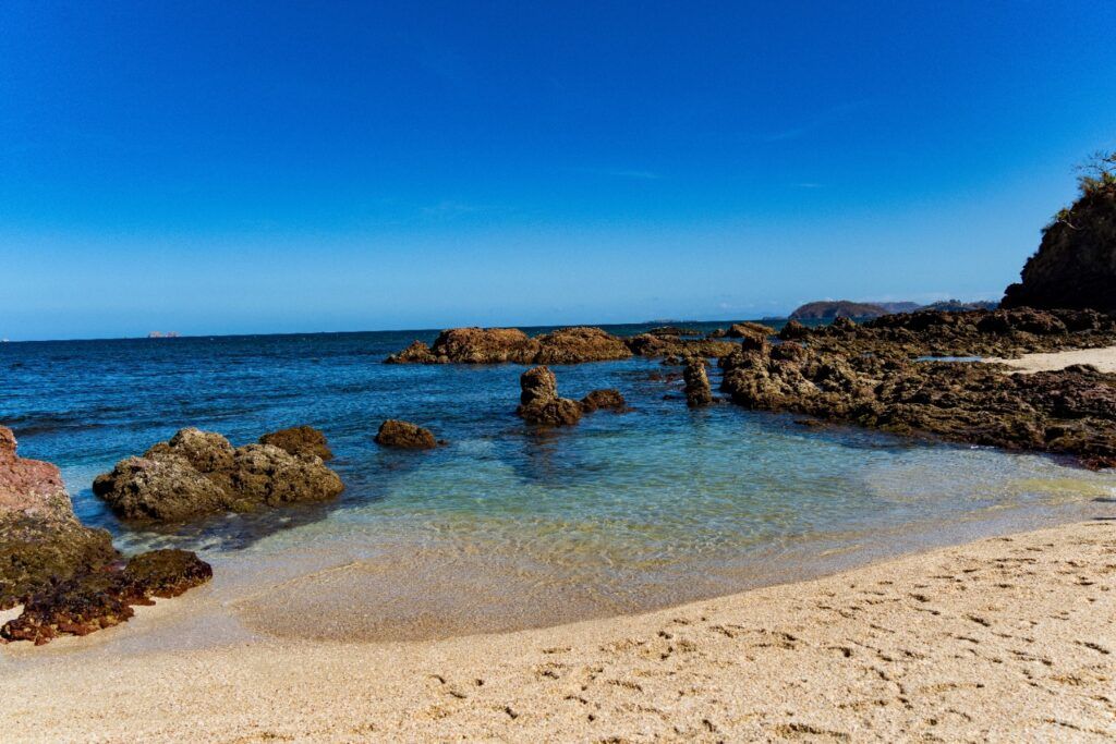 Sandy beach with clear shallow water, rocky outcrops, and dark blue ocean under a bright blue sky.
