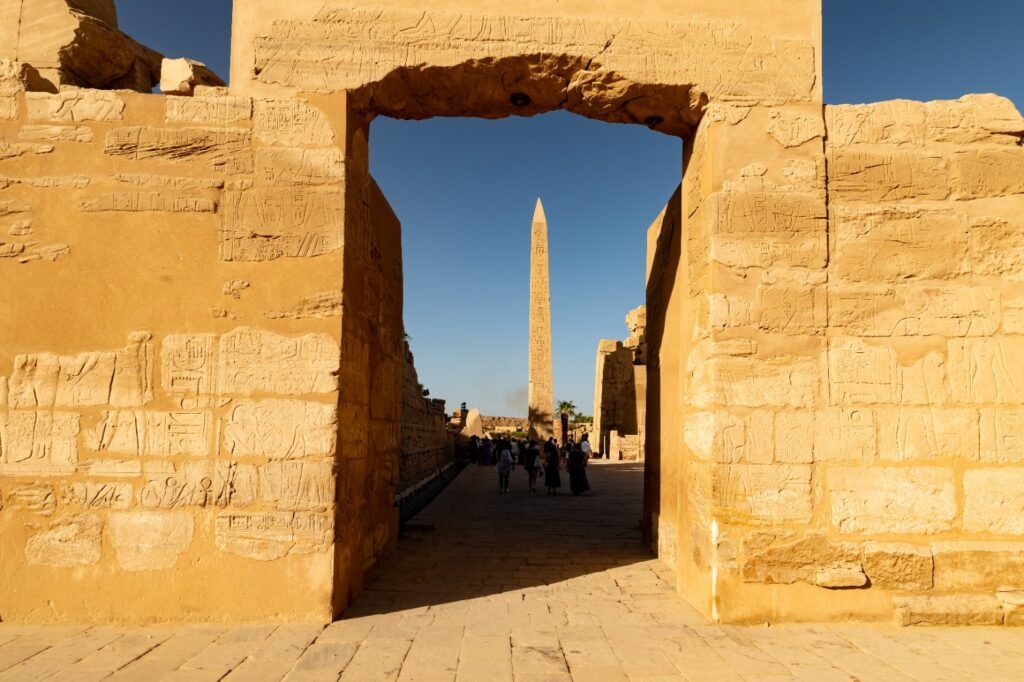 Ornate, inscribed stone archway framing a distant view of a tall Egyptian obelisk under a bright blue sky, with a group of tourists walking below at the Karnak Temple.