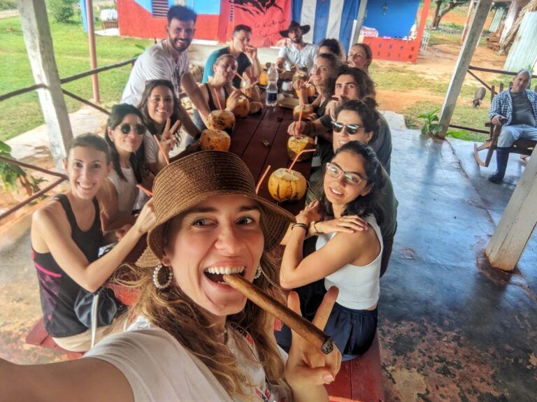 A group of smiling WeRoad travelers enjoying fresh coconuts and cigars together at a long wooden table in Cuba.
