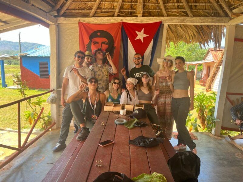 A group of WeRoad travelers posing with cigars in front of a Cuban flag and a portrait of Che Guevara in an outdoor hut.