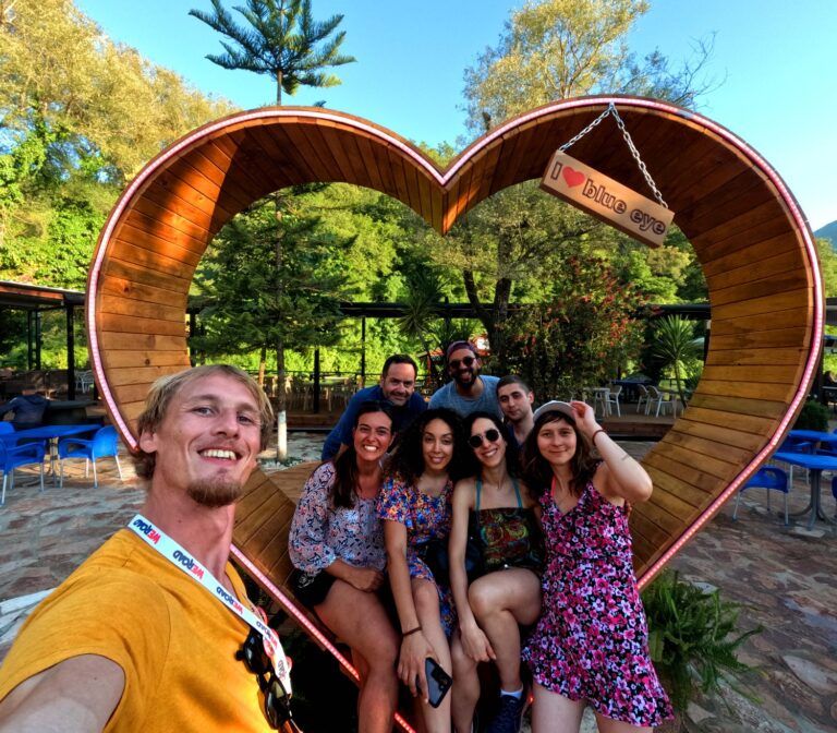 A smiling group of WeRoad travelers posing inside a large wooden heart-shaped frame at the Blue Eye in Albania.