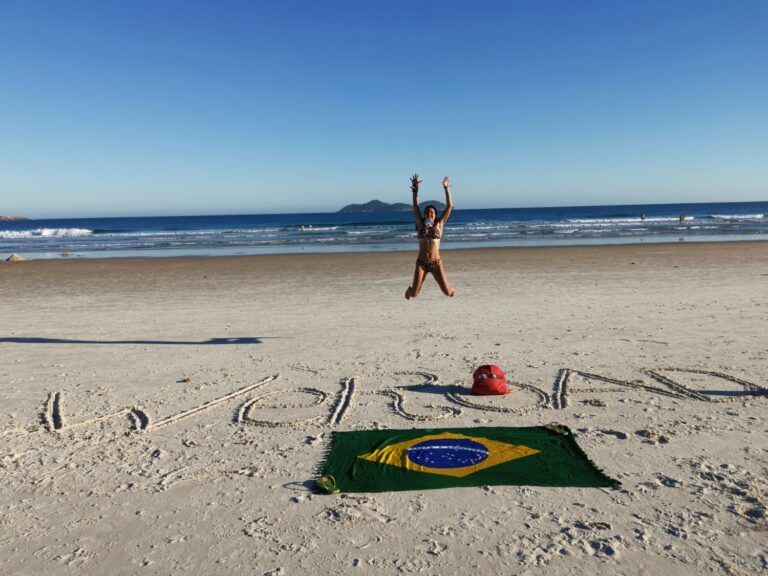A traveler jumping for joy on a white sand beach in Brazil, with 'WeRoad' written in the sand and a Brazilian flag spread out.