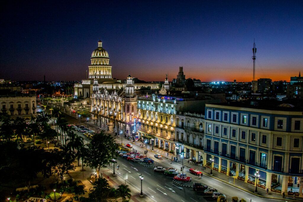 A high-angle nighttime view of a wide street in Havana, Cuba, featuring the illuminated National Capitol Building and ornate historic hotels under a deep blue twilight sky.