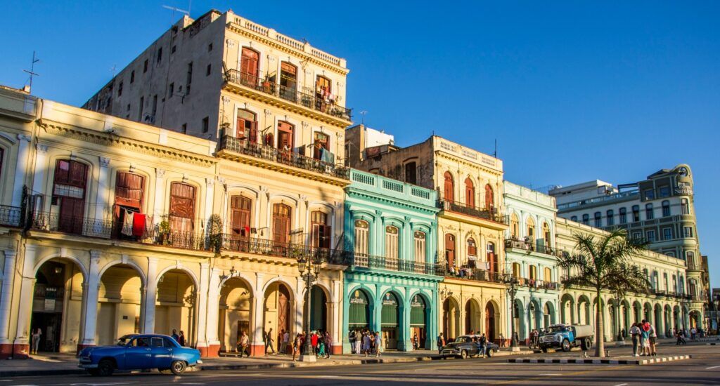 A row of vibrant, colorful historic buildings in Havana, Cuba, with ornate balconies and arched walkways, under a clear blue sky with a classic blue car parked in the foreground.