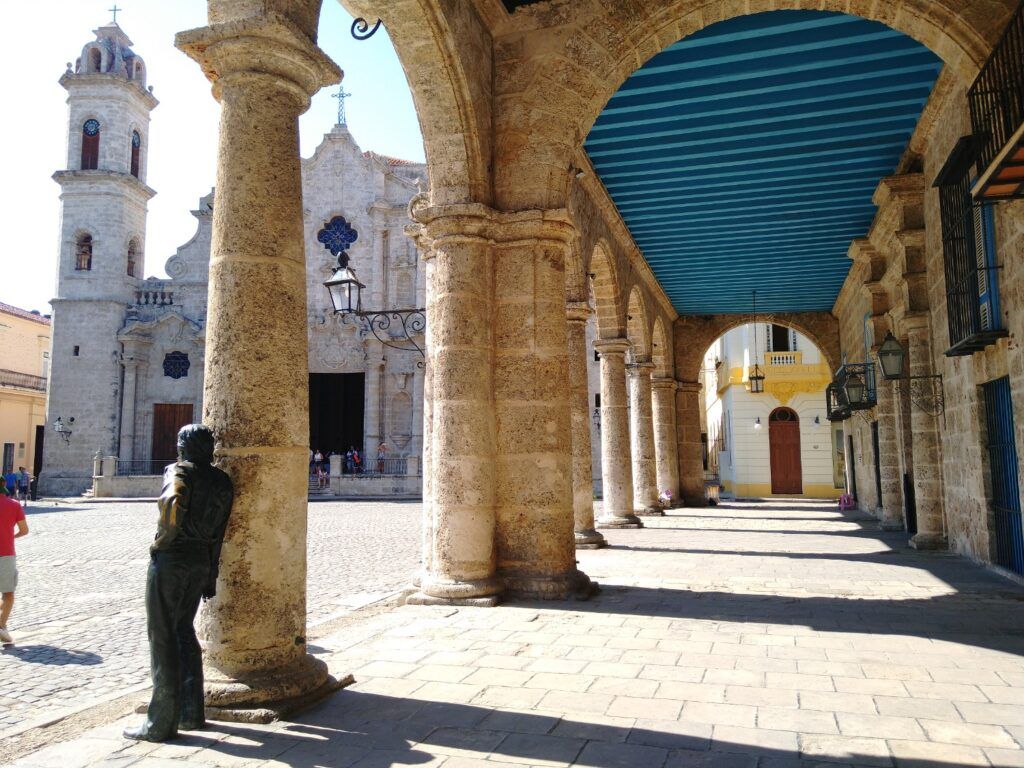 View of Havana Cathedral through a series of stone arches, with a bronze statue of a man leaning against a pillar in the foreground.