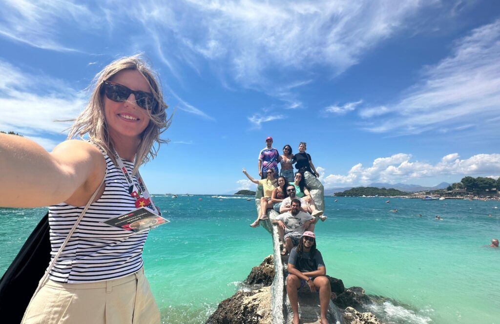 A group of WeRoad travelers posing on a hand-shaped stone sculpture at a turquoise beach in Ksamil, Albania.