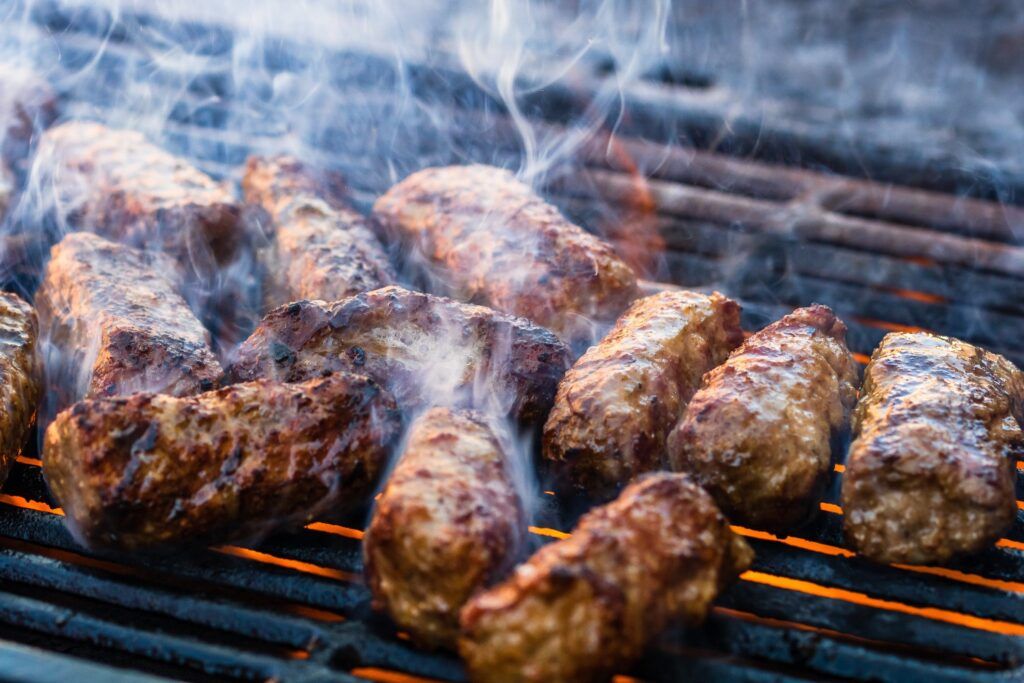 Close-up of several Mici, traditional Romanian grilled ground meat rolls, cooking on a hot grill with smoke rising.