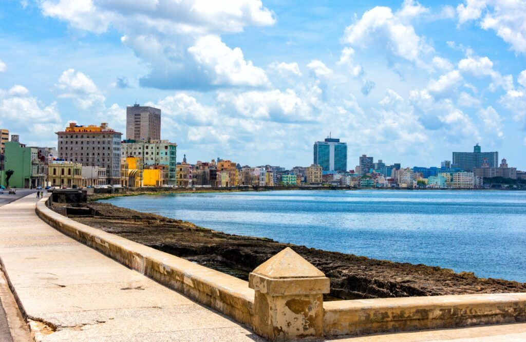 A wide view of the Malecón waterfront in Havana, Cuba, showing a curved stone promenade alongside the blue sea with the city skyline and colorful buildings under a cloudy sky.