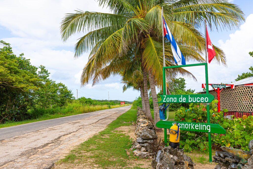 A roadside sign marking a diving and snorkeling zone next to palm trees and a Cuban flag.