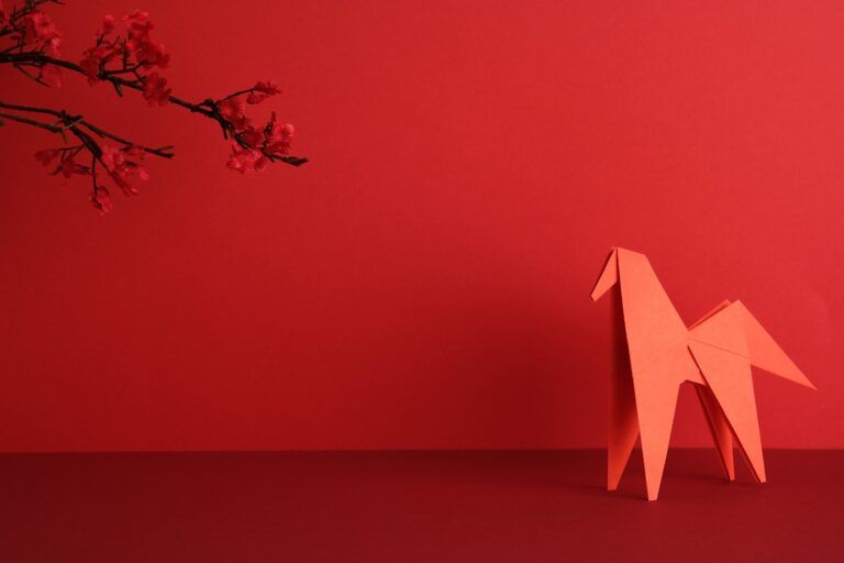 A red origami horse placed next to a cherry blossom branch on a red background, celebrating the Year of the Horse for the 2026 Chinese New Year.