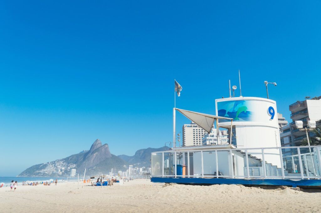 Ipanema Beach in Rio de Janeiro, featuring a white lifeguard station with the number 9 and Two Brothers Hill in the distance.