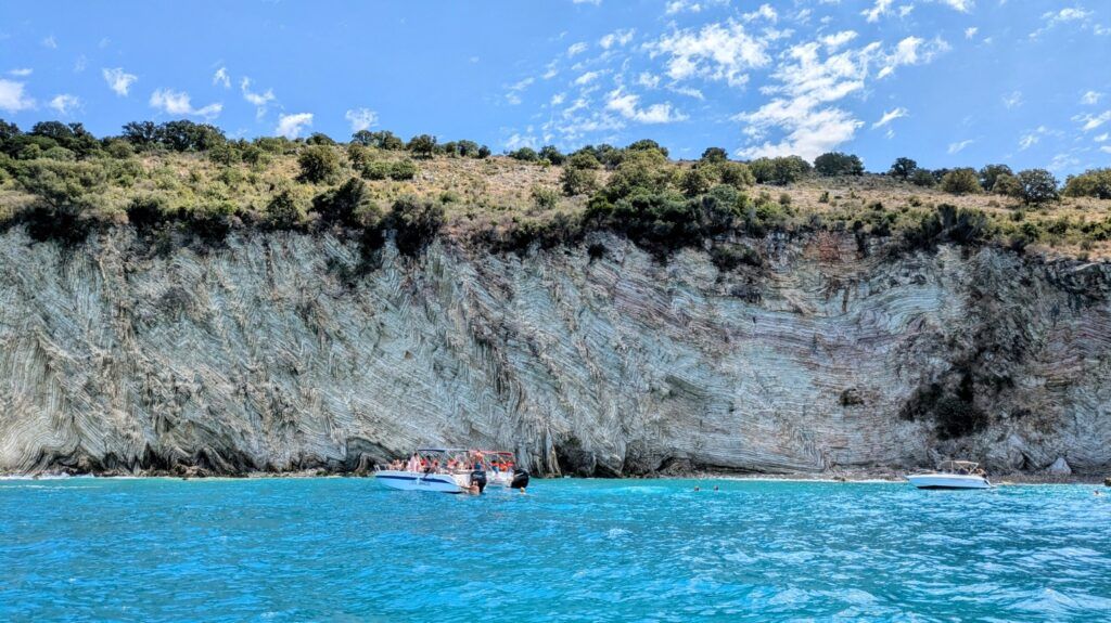 Tour boats floating on bright turquoise water next to a massive, unique striped limestone cliff in Albania.
