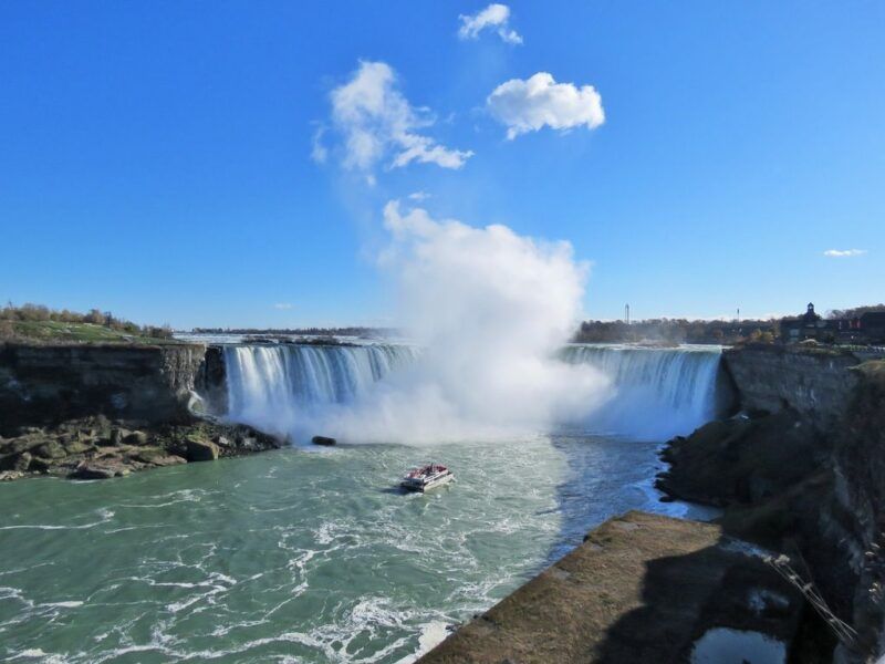 A powerful panoramic view of the Horseshoe Falls at Niagara Falls, Canada, with a tour boat approaching the mist under a clear blue sky.