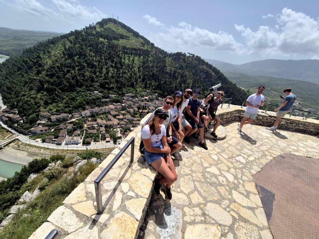 A group of WeRoad travelers sitting on a stone wall at a panoramic viewpoint overlooking the historic city of Berat, Albania, and its green hills.